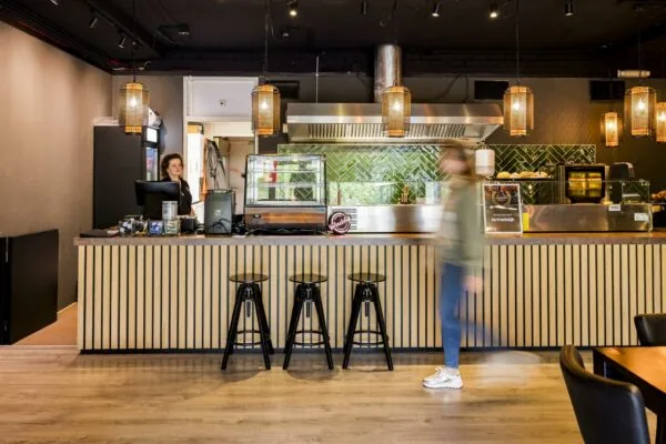 Modern ingerichte horecagelegenheid aan de Markerkant met houten bar, hanglampen en een medewerker achter de kassa.